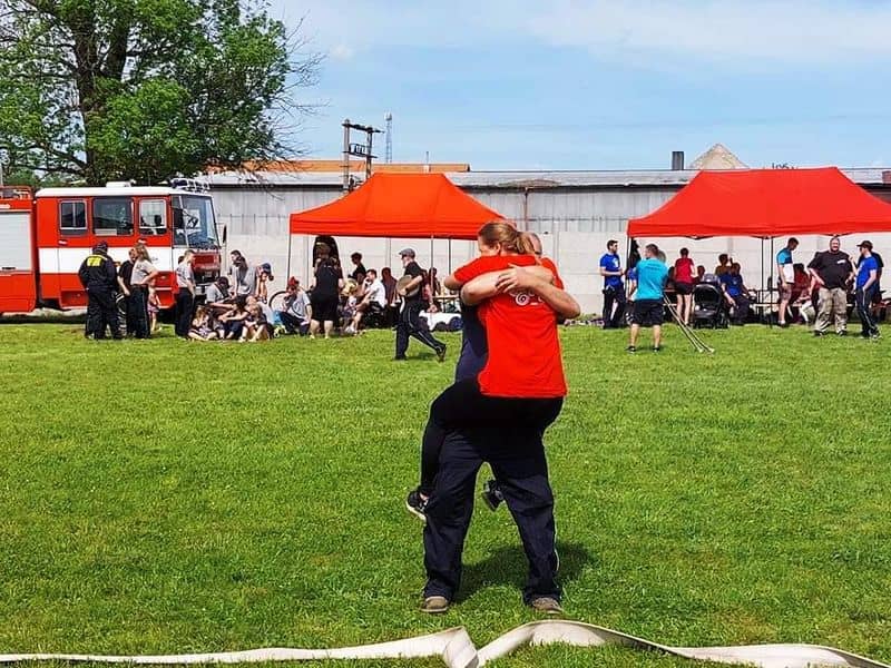 Dva lidé se objímají na travnaté ploše, v pozadí hasičské auto a stany