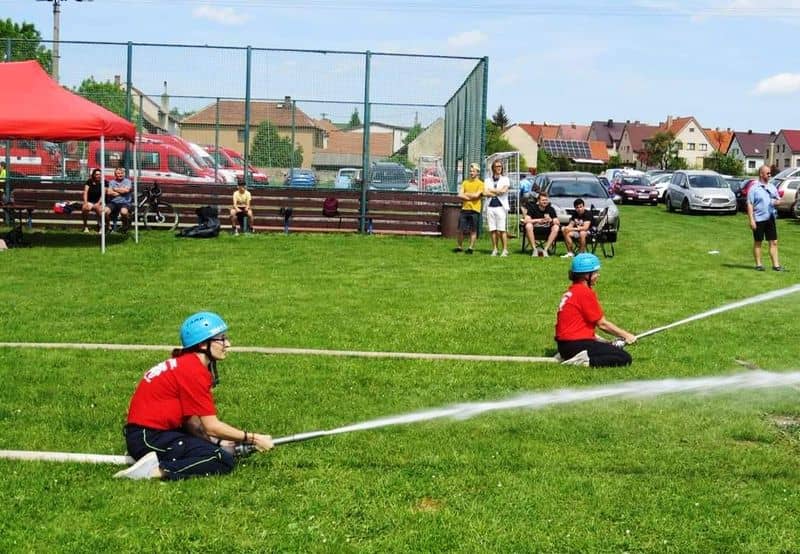 Dvě ženy v hasičských uniformách stříkají vodu z hadic na travnatém hřišti