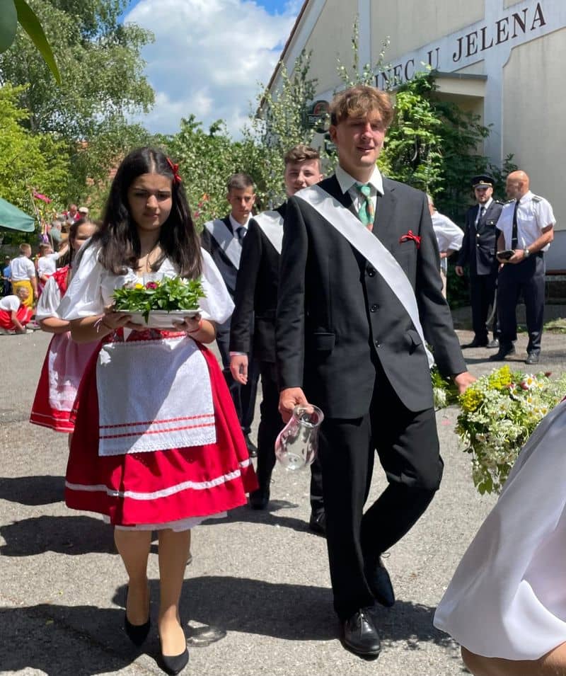Dívka v kroji s květinami a muž v obleku s šerpou a džbánem