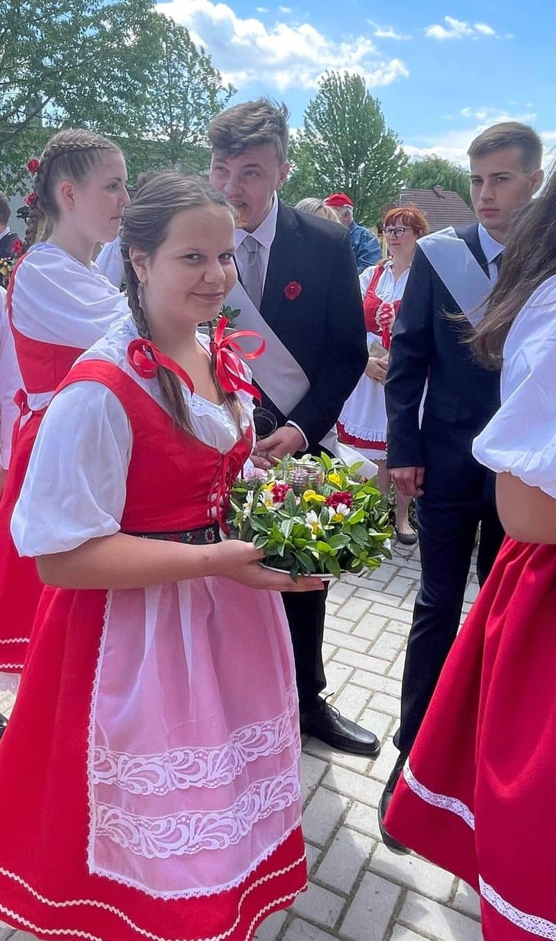 Dívka v kroji drží věnec s květinami, za ní skupina mladých lidí