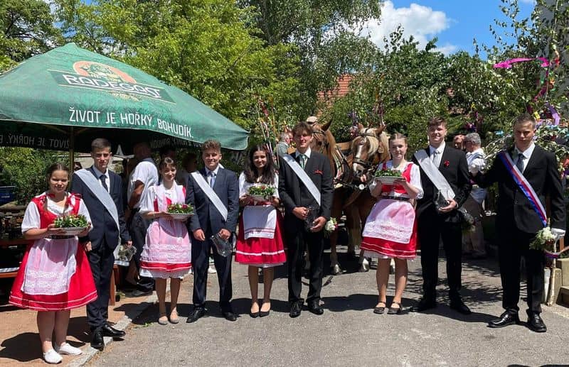 Skupina mladých lidí v krojích a oblecích s šerpami, za nimi koně