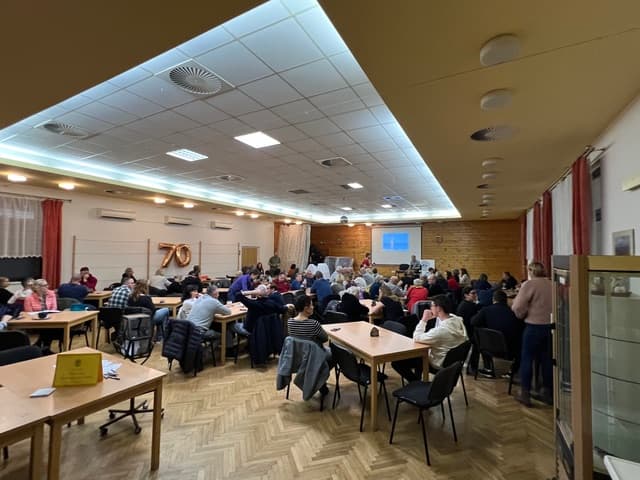 Místnost plná lidí sedících u stolů, na stěně číslo 70