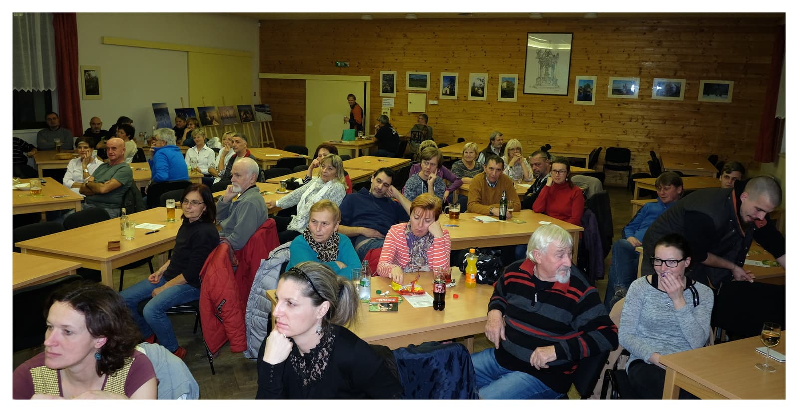 Skupina lidí sedí u stolů v sále s dřevěným obložením