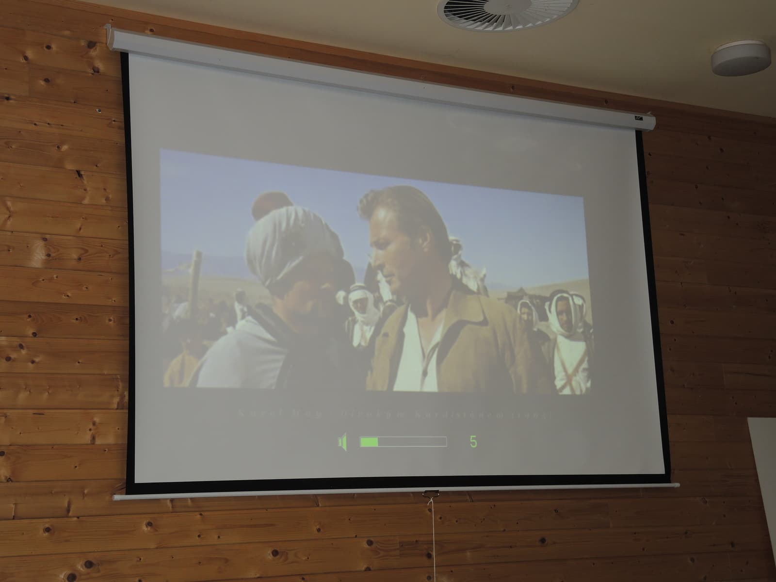 Promítání filmu s pouštní scénou na plátně v sále s dřevěným obložením