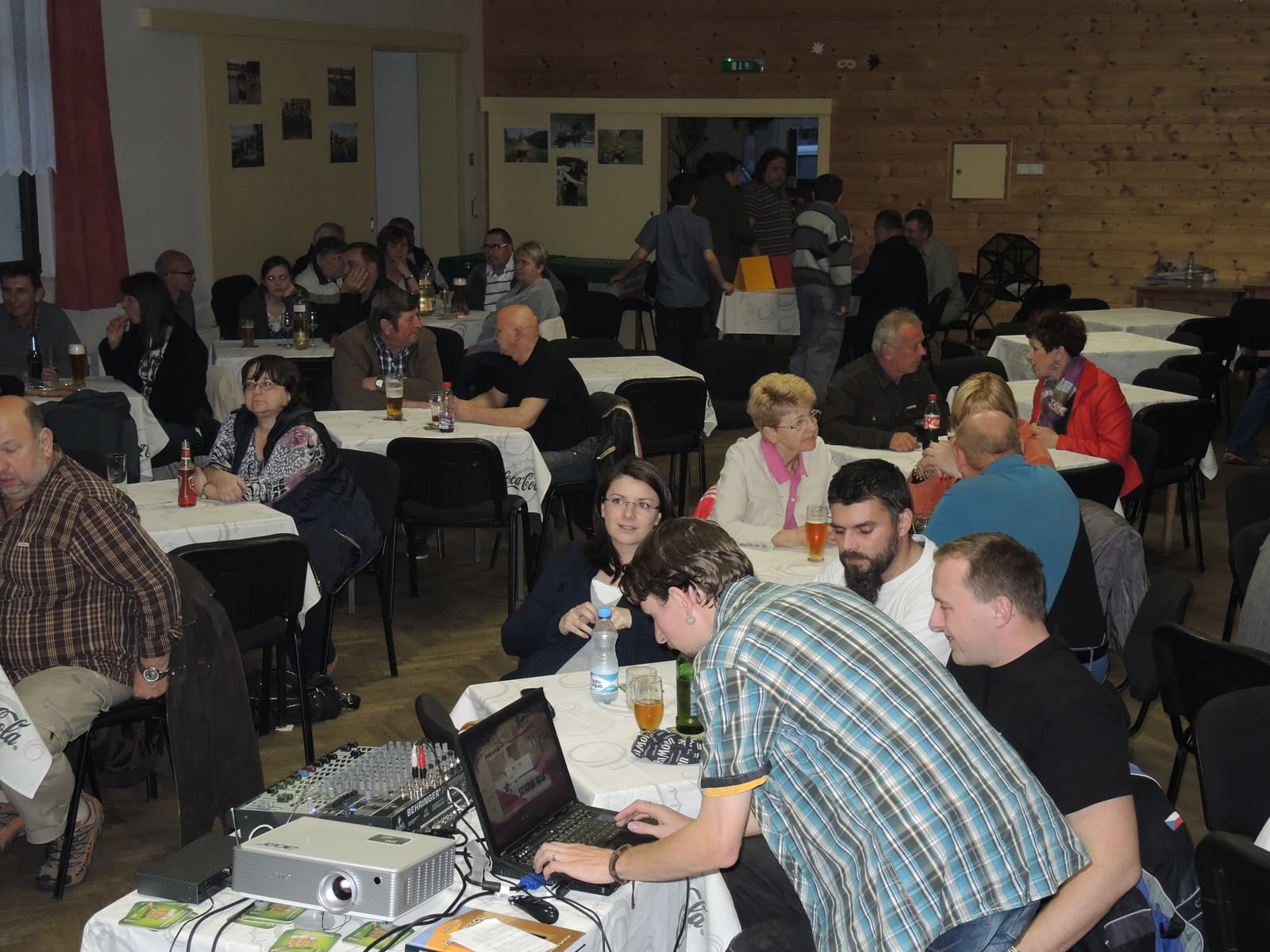 Skupina lidí sedí u stolů v sále, v popředí muž u notebooku a projektoru