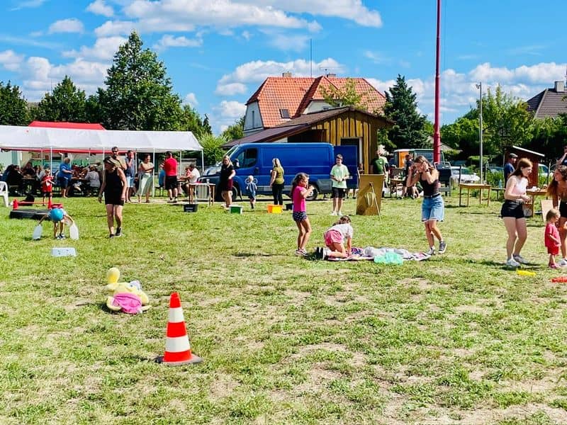 Skupina lidí na venkovní akci, děti si hrají na trávě
