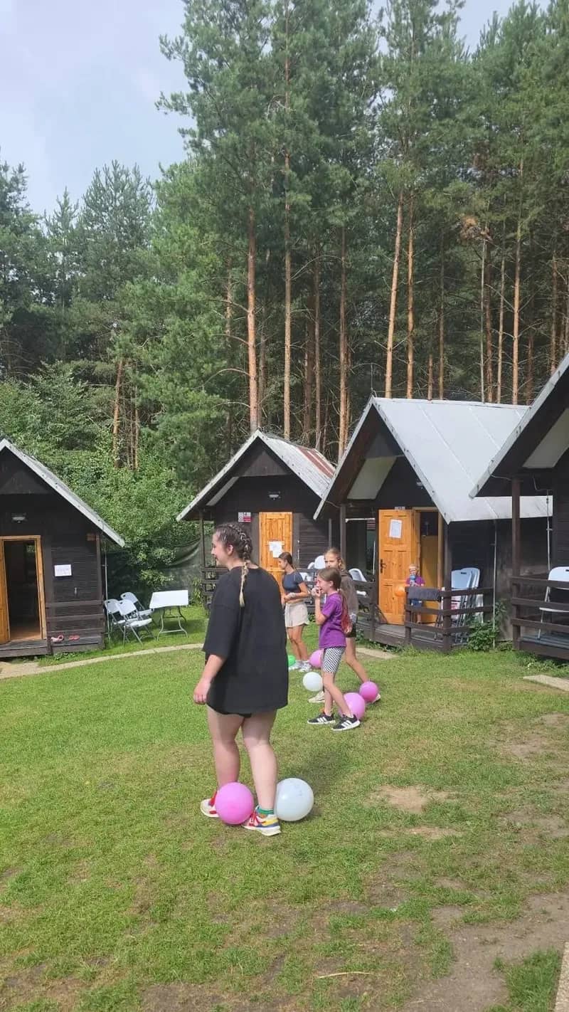 Dvojice dětí s balónky před táborovými chatkami.