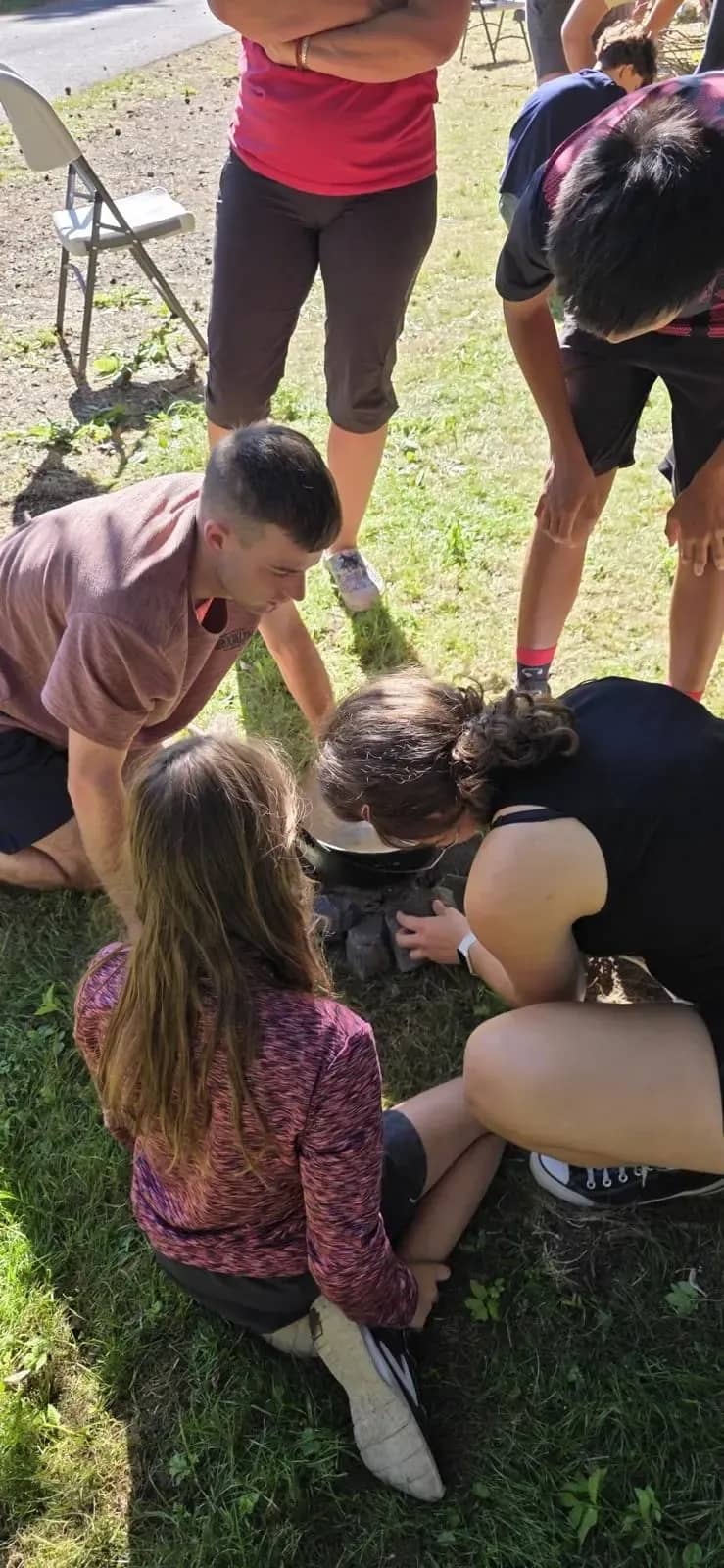 Skupina se sklání nad surovinami při přípravě jídla venku.