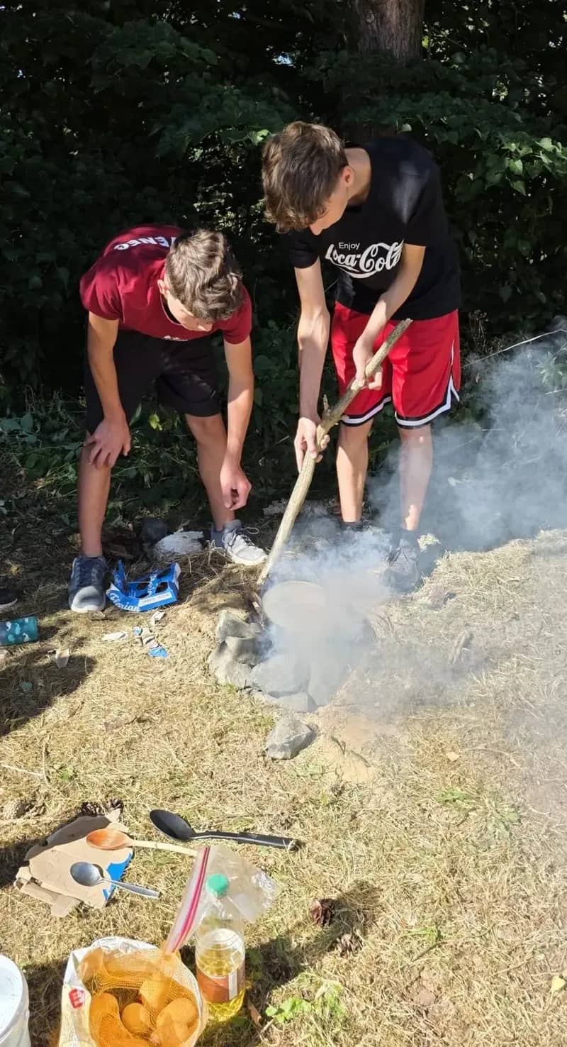 Účastníci připravují jídlo u kouřícího ohniště.