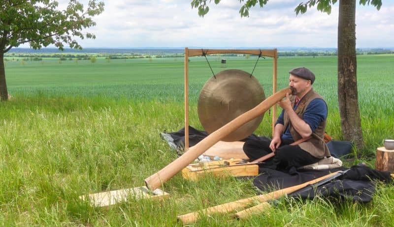Muž hraje na didgeridoo a gong v trávě pod stromem