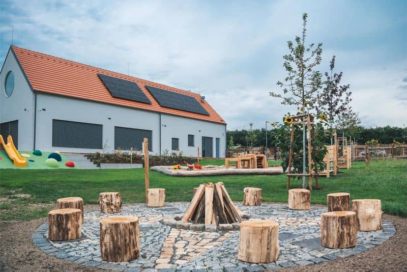 Nová budova s hřištěm, pískovištěm a ohništěm s pařezy k sezení