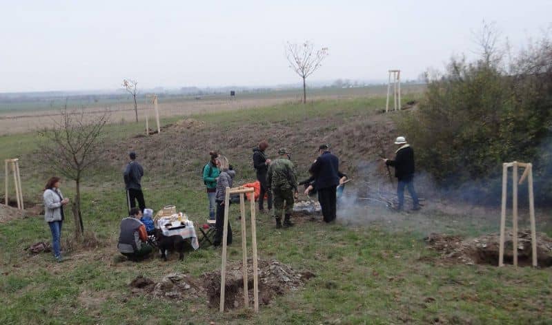 Skupina lidí u ohně a stolu v krajině s nově zasazenými stromy
