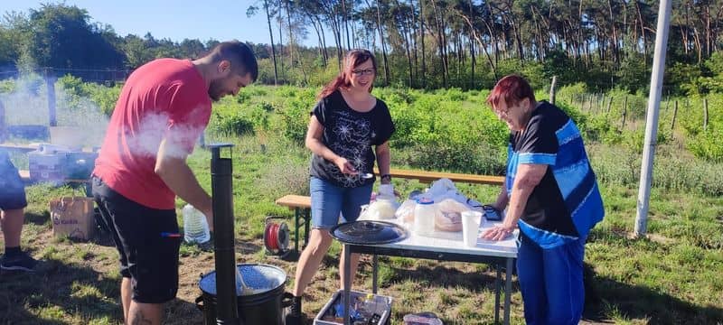 Tři lidé připravují jídlo venku, muž míchá v kotli, dvě ženy stojí u stolu