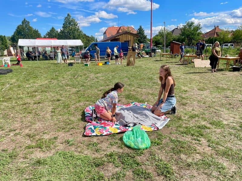 Dvě dívky na dece na trávě, v pozadí skupina lidí a stany