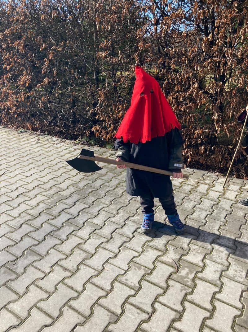 Dítě v červené kápi s otvory pro oči drží sekeru