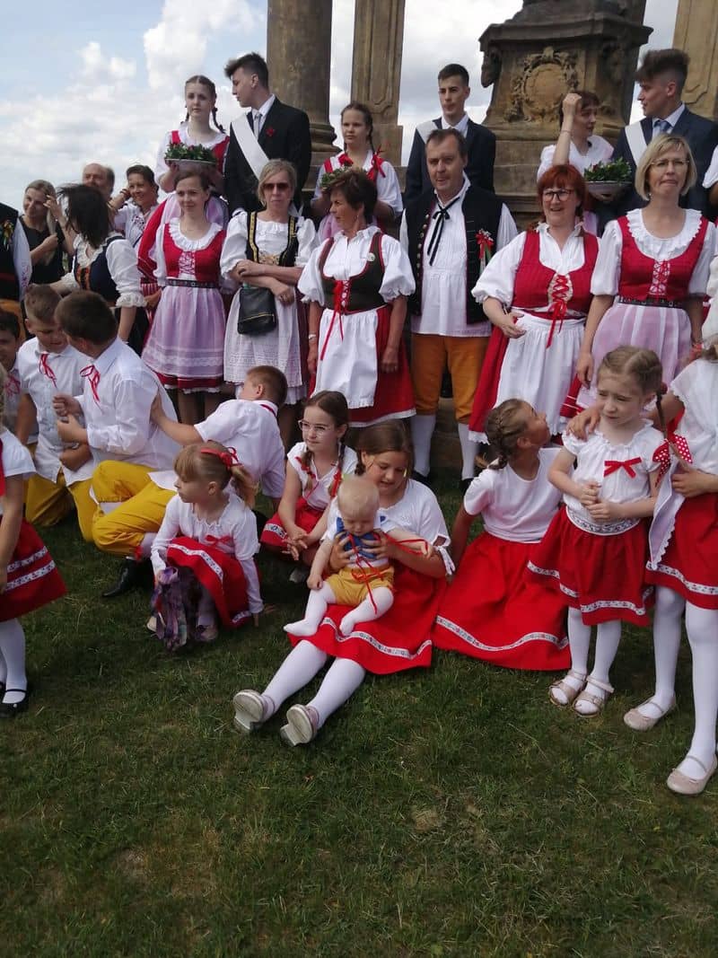 Skupina lidí v krojích, dospělí stojí, děti sedí na trávě