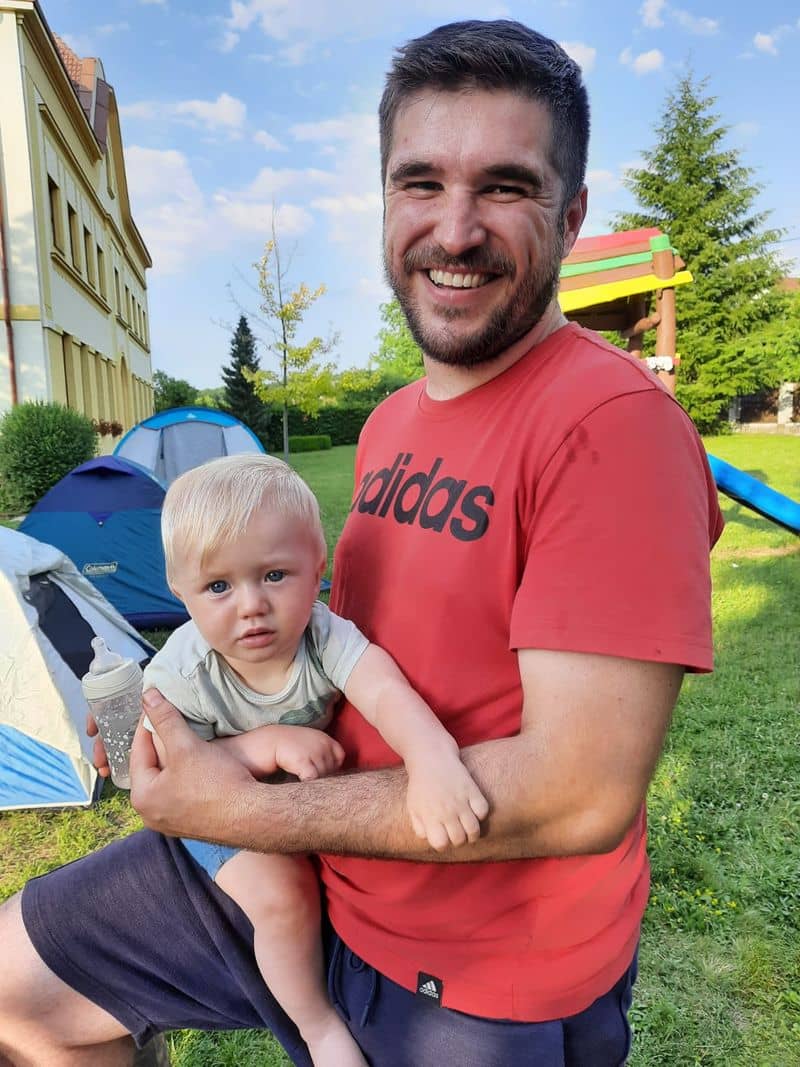 Muž drží dítě, v pozadí stany a budova