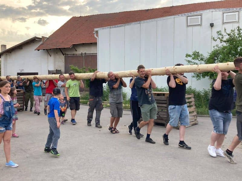 Skupina mužů nese dlouhou oloupanou kládu, kolem nich jdou děti