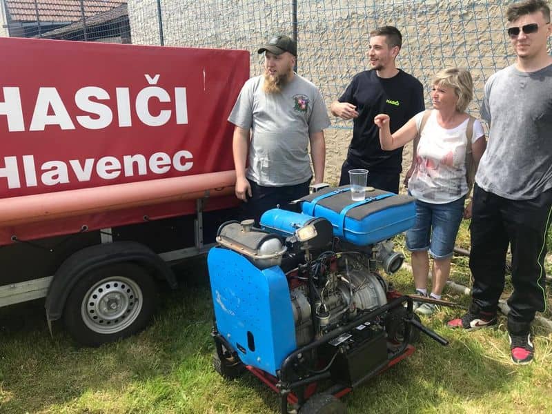 Skupina lidí u hasičského přívěsu s nápisem Hasiči Hlavenec a modré stříkačky