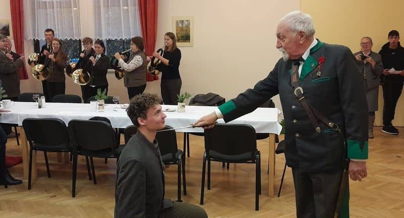 Muž v kroji pasuje mladíka mečem, v pozadí hraje dechová kapela