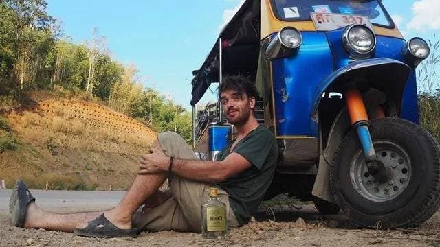 Muž sedí u tuk-tuku s lahví alkoholu, v pozadí kopec
