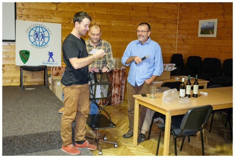 Tři muži na pódiu, jeden drží mikrofon, druhý čte z papíru