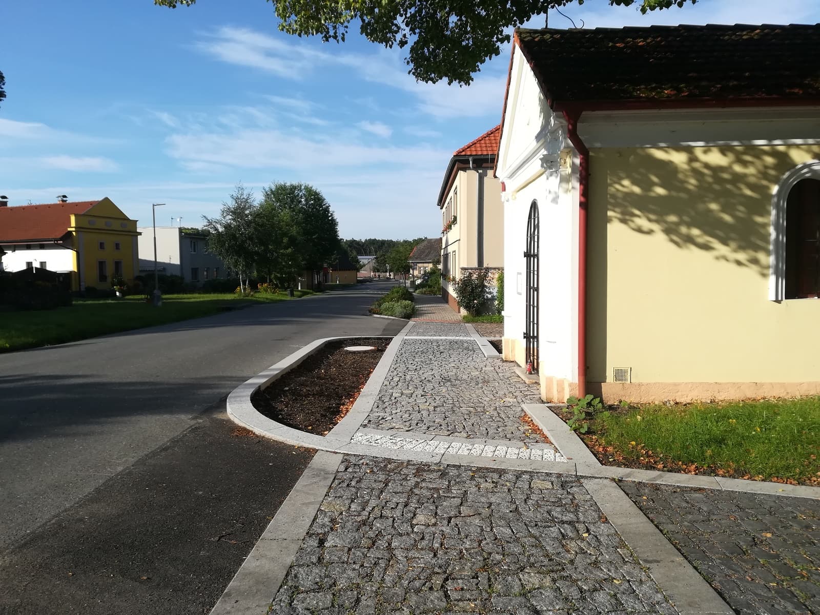 Dlážděný chodník a silnice v obci, vpravo kaplička