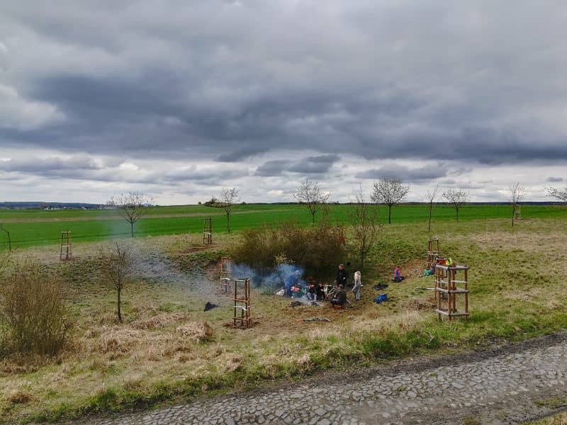Skupina lidí u ohně v krajině s mladými stromy pod zataženou oblohou