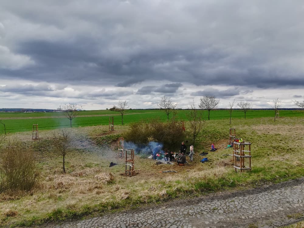 Skupina lidí u ohně v krajině s mladými stromy pod zataženou oblohou