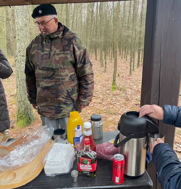 Muž v maskáčové bundě a baretu u stolu s jídlem a pitím v lese