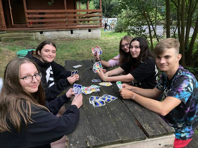 Skupina pěti mladých lidí hraje karty u dřevěného stolu venku