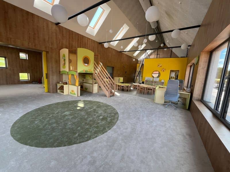 Herna mateřské školy s vysokým stropem, střešními okny, kulatým kobercem a dřevěným domečkem na hraní