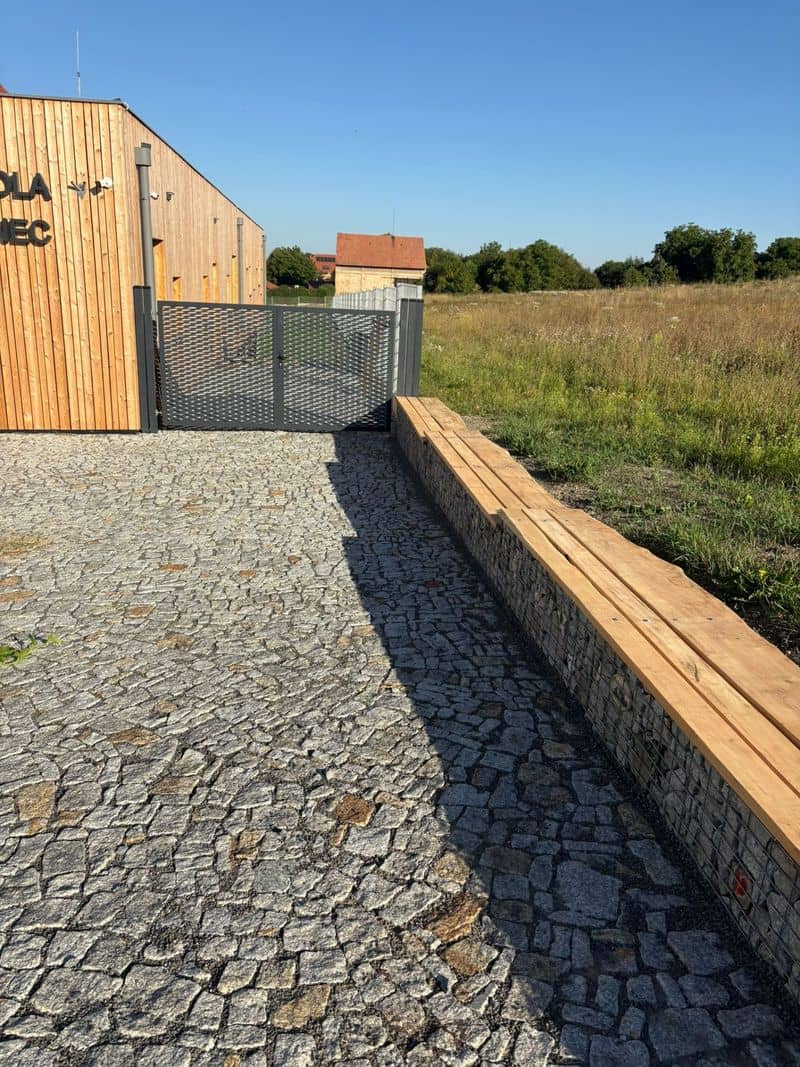 Dlážděný vstup k mateřské škole s brankou do zahrady, za plotem pole