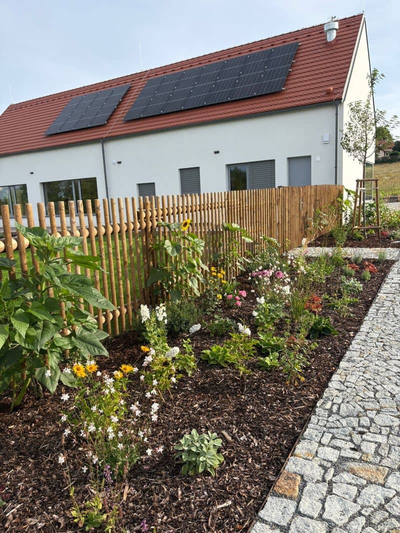 Zahradní záhon s květinami u dřevěného plotu, v pozadí budova se solárními panely