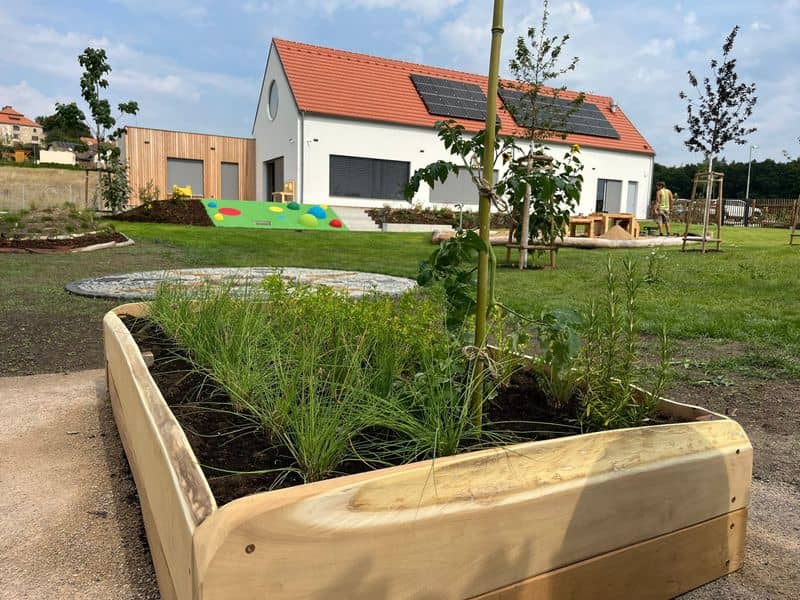 Budova mateřské školy zvenku, na zahradě vyvýšený záhon s bylinkami a nově vysázené stromy