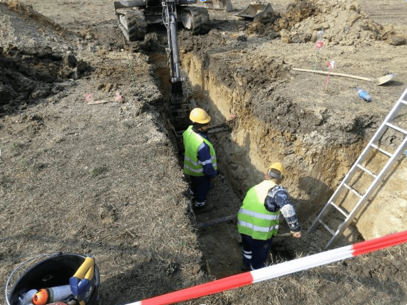 Dva muži v reflexních vestách pracují v hlubokém výkopu