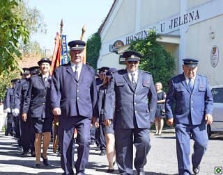 Skupina hasičů v uniformách pochoduje před budovou Hostince U Jelena