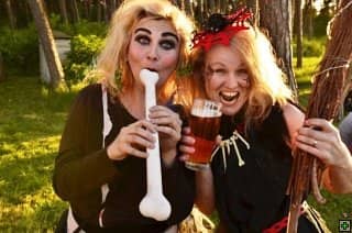 Dvě ženy v kostýmech čarodějnic, jedna drží píšťalu ve tvaru kosti, druhá pivo