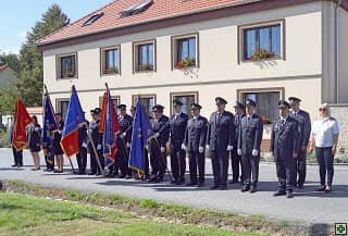 Skupina hasičů v uniformách s prapory před budovou