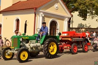 Muž v modré kombinéze řídí zelený historický traktor s přívěsem