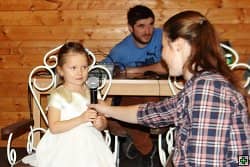 Žena podává mikrofon malé holčičce v bílých šatech, v pozadí muž