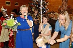 Žena s mikrofonem a kyticí, děti a další lidé u vánočního stromku