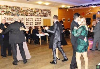 Skupina lidí tančí v sále, v pozadí stoly a nástěnka s fotografiemi