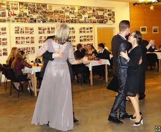 Pár tančí v sále, v pozadí další lidé u stolů a stěna s fotografiemi