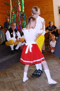 Děti v krojích tančí, v pozadí sedí dospělí