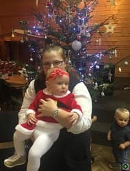 Žena drží dítě v kostýmu Santa Clause, v pozadí vánoční stromek a další dítě
