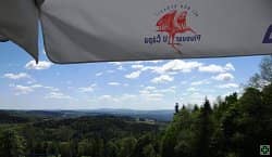 Krajina s lesy a kopci pod modrou oblohou, v popředí část slunečníku s logem