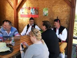 Skupina lidí sedí u stolu pod dřevěným přístřeškem, dva muži v krojích