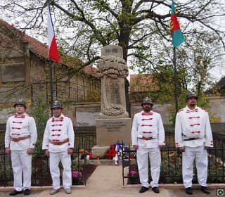 Čtyři muži v historických uniformách u pomníku padlých s českou a obecní vlajkou