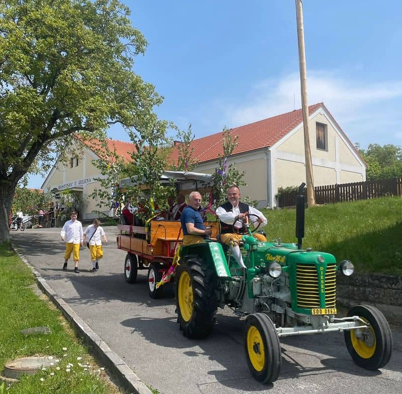 Muži v krojích na traktoru s vlekem a děti v krojích na cestě